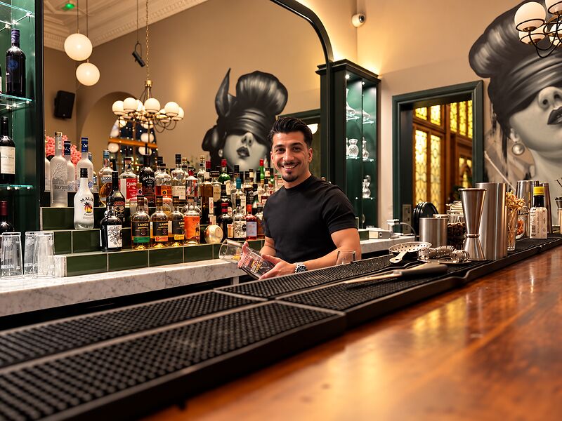 A smiling bartender behind the polished timber bar, with the mural and bottle shelves behind.