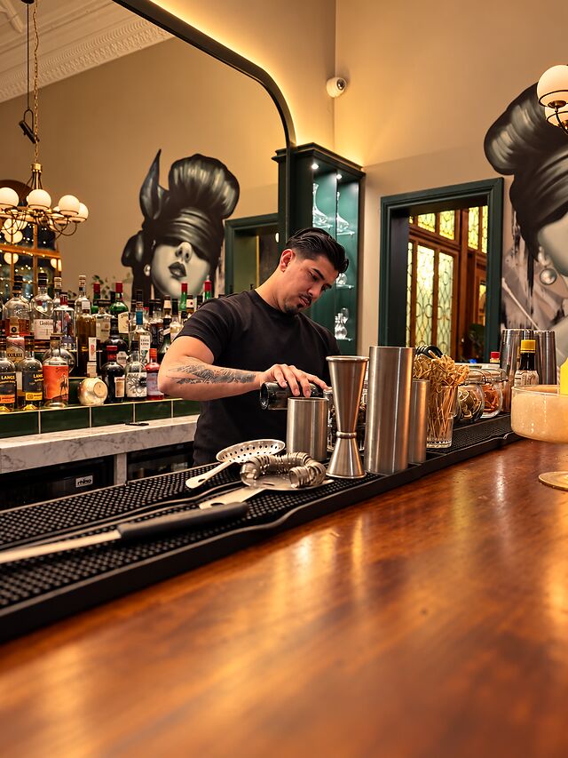 A bartender pouring a drink at the polished timber bar, with the mural and bottle shelves behind.