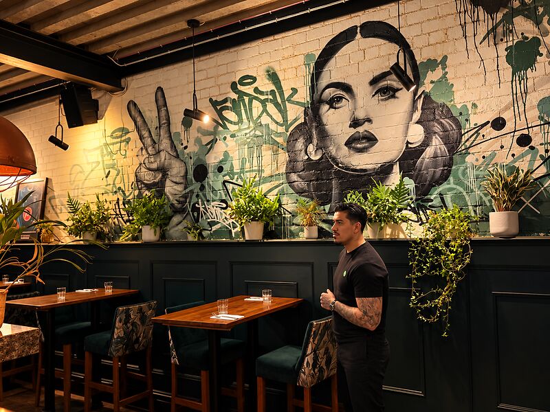 A member of the team standing in front of a street-art mural of a woman's face, beside timber tables and green stools.